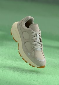 Beige träningsskor med en texturerad ovandel, snörning och rundad sula. Har en ljusblå accent och en orange gummisula.