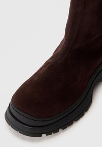 Rudos zomšinės aulinės suapvalintu noseliu ir storu juodos gumos padu, viršutinėje briaunoje ir padai išryškintais tekstūruotais elementais.