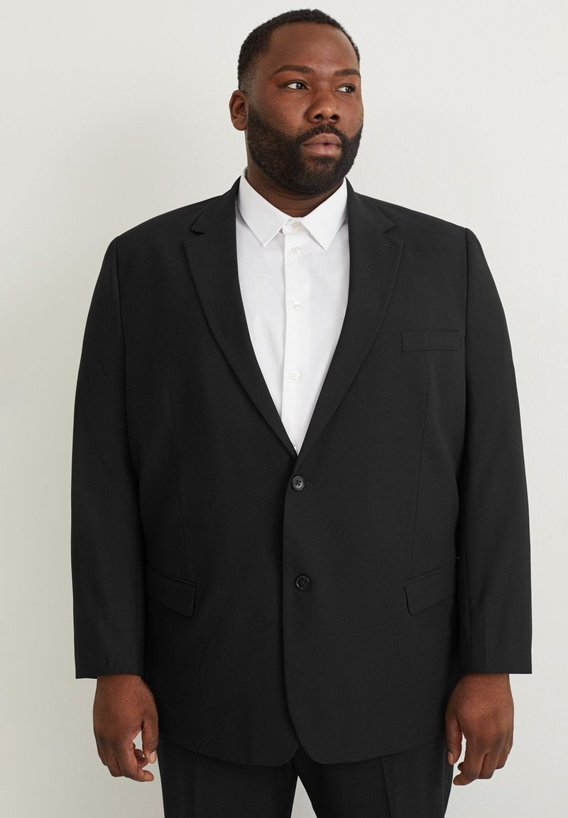 Blazer negro con dos botones, solapas con muesca y un bolsillo en el pecho, combinado con una camisa blanca abotonada. Textura de tela suave.