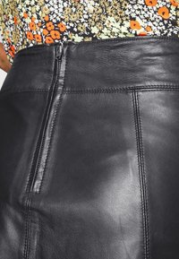 Svart läderskjorta med en slät yta, utrustad med en bakre dragkedja och sömdetaljer. Midjebandet matchar kjolens material.