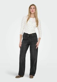 Haut blanc à manches longues avec des pois, ourlet festonné. Jeans noirs taille haute à jambes larges. Mannequin portant des chaussures plates marron.