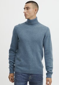 Pull à col roulé bleu en tissu doux, avec un col et des poignets côtelés, de coupe standard, associé à un jean sombre.