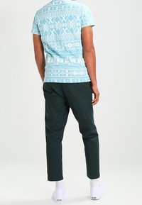 Camiseta de manga corta azul claro con patrones geométricos blancos en la parte de atrás, combinada con pantalones oscuros verdes y zapatillas blancas.