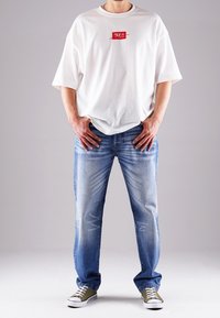 Biała, oversize'owa koszulka z czerwonym logo graficznym, zestawiona z jasnoniebieskimi jeansami z efektami przetarć i detalami szwów, noszona z zielonymi trampkami.
