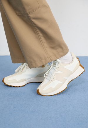 Hvide og beige sneakers med gummisåler båret med hvide sokker og beige bukser på et blåt tæppebelagt gulv.