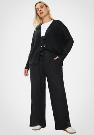 Femme aux cheveux blonds portant un cardigan noir sur un haut blanc, un pantalon large noir et des baskets blanches, debout avec une main dans la poche.