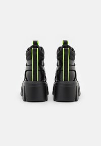 Baskets noires avec une semelle épaisse et un talon surélevé. Présentent des coutures vert vif le long du talon et une languette à l'arrière.