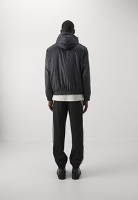 Hombre de pie de espaldas, vistiendo una chaqueta con capucha negra, una camisa blanca debajo, pantalones negros y zapatos negros, contra un fondo claro y sencillo.