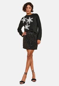 Melnā džemperis ar balto sniegpārsliņu dizainu, kombinēts ar melnu mini svārku ar paildzinātām detaļām un melnu ziedu jostu ar augstiem papēžiem. Gludas tekstūras, pieguļošs stils.