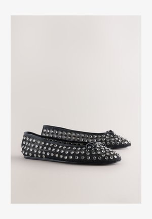 Schwarze flache Schuhe mit spitz zulaufender Spitze, verziert mit mehreren runden silbernen Nieten und einem Schleifenakzent vorne. Glatte Textur.