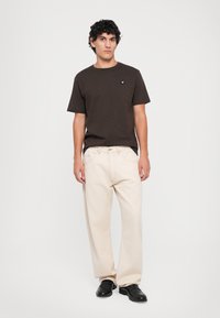 Junger Mann mit lockigem, dunklem Haar trägt ein dunkelbraunes T-Shirt, helle, weit geschnittene Hose und schwarze Schuhe und steht vor einem weißen Hintergrund.