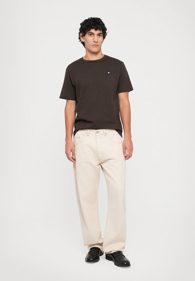 Un jeune homme aux cheveux bouclés et foncés porte un t-shirt marron foncé, un pantalon crème à jambes larges et des chaussures noires, se tenant devant un fond blanc.