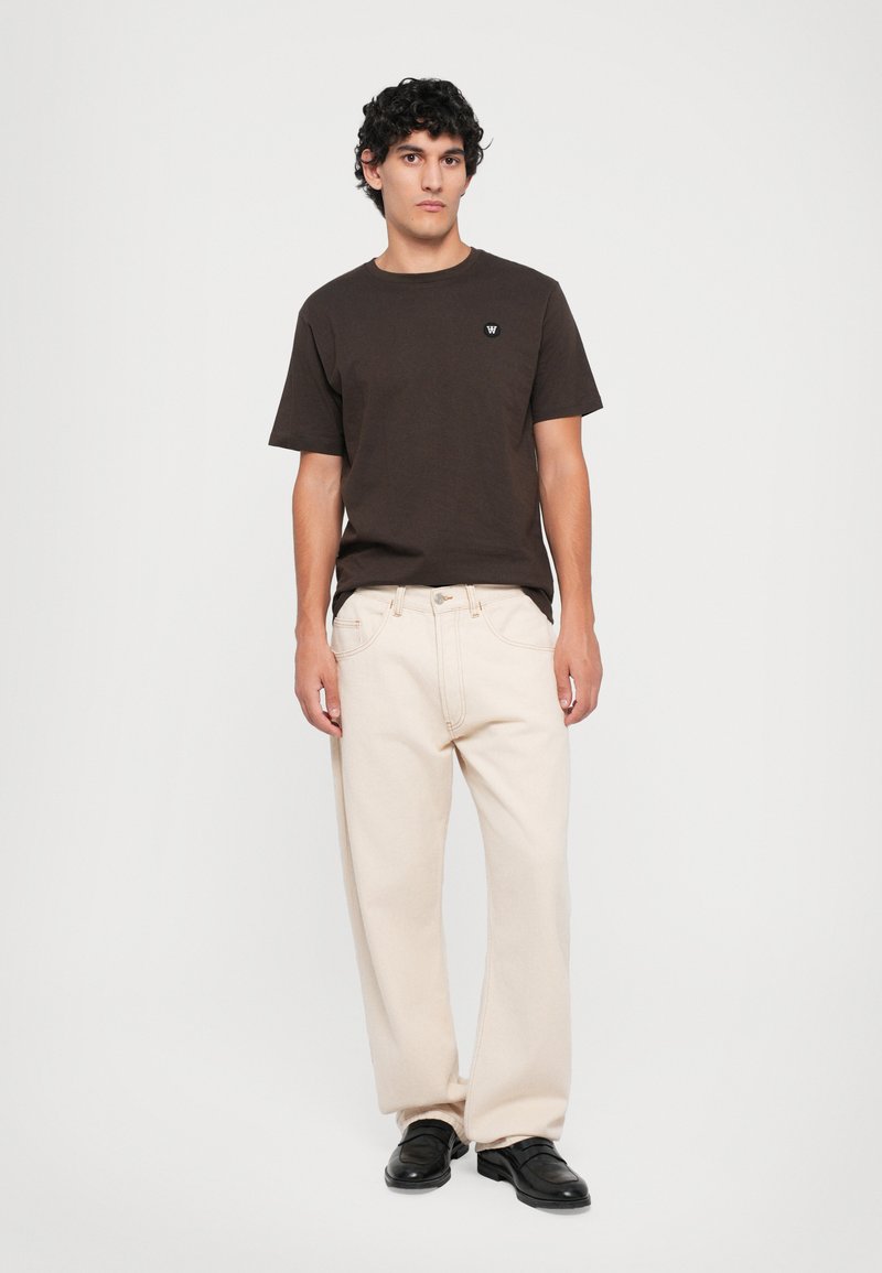 Junger Mann mit lockigem, dunklem Haar trägt ein dunkelbraunes T-Shirt, helle, weit geschnittene Hose und schwarze Schuhe und steht vor einem weißen Hintergrund.