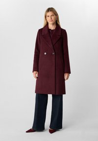 Donna che indossa un lungo cappotto bordeaux doppiopetto con grandi revers, pantaloni larghi scuri e scarpe a punta bordeaux coordinate, in piedi.