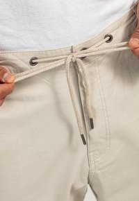 Beige Shorts mit einem Stoffkordelzug, Metallösen, strukturierter Oberfläche und cremefarbenen Nähten, gehalten an der Taille von Händen.