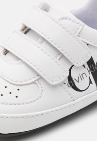 Białe skórzane trampki z rzepami, perforowanymi detalami na palcach oraz czarnym logo Calvin Klein z boku. Płaska podeszwa dla stabilności.