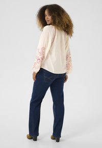 Crèmekleurige blouse met rode geborduurde bloemenpatronen op de mouwen, gecombineerd met een blauwe spijkerbroek. Het model heeft natuurlijk krullend haar en staat met de rug naar de camera.