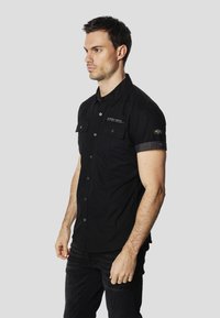 Camisa negra de manga corta hecha de tela con textura suave; cuenta con dos bolsillos delanteros, cierre de botones y detalles de logotipo en el pecho y la manga.