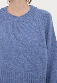 Gros plan sur une personne portant un pull en tricot doux bleu avec un col rond côtelé et des manches raglan.