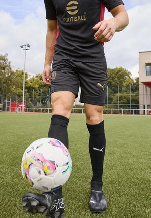 Svart fotbollsklädsel med guldaccenter, svarta strumpor och sportskor. Färgglad mönstrad fotboll rullar framför på konstgräs.