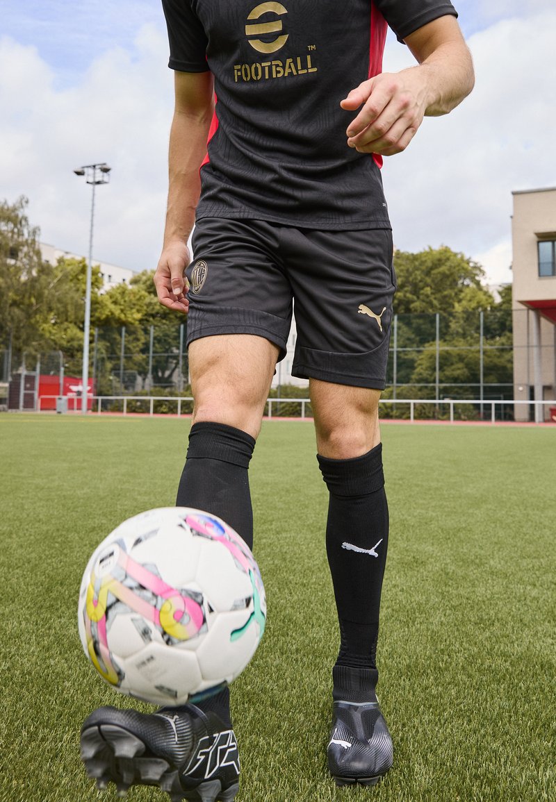 Svart fotbollsklädsel med guldaccenter, svarta strumpor och sportskor. Färgglad mönstrad fotboll rullar framför på konstgräs.