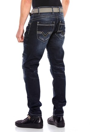 Dunkelblaue Jeans aus Denim mit Slim-Fit, abgewetzten Details, weißen Nähten und einzigartigem Taschendesign. Getragen mit einem strukturierten grauen Gürtel und schwarzen Schuhen.