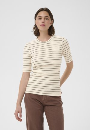 Femme aux cheveux courts et foncés portant un haut côtelé crème à manches courtes avec de fines rayures horizontales marron et un pantalon marron, debout devant un fond uni.