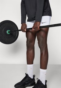 Svart långärmad träningsskjorta, vita shorts med Nike-logotyp, greppande en skivstång med svarta gummivikter. Svarta sneakers med vita strumpor.