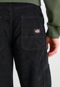Mustat corduroy-housut, joissa on uritettu pinta, suuret takataskut ja Dickies-logo-lappu vyötäröllä.