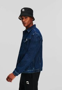 Donkerblauwe spijkerjack met metalen knopen, borstzak en witte logo patch. Zwarte bucket hat met logo, gedragen boven zwarte broek.