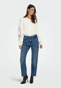Blusa blanca de manga larga con detalles de encaje en las mangas, combinada con jeans de mezclilla azules de cintura alta y botas negras brillantes hasta el tobillo.