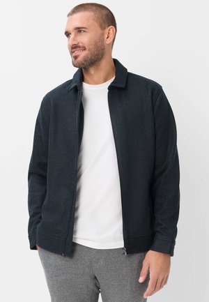 Mand iført en mørkeblå lynlåstrøje over en hvid t-shirt og grå bukser, kigger til siden med hænderne i lommerne.