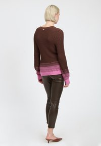 Vrouw met kort blond haar, gekleed in een gestructureerde bruine trui met roze boorden en zoom, donkere leren broek en bruine hakken, met haar rug naar ons toegekeerd.