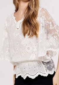 Blusa blanca bordada con mangas murciélago transparentes, dobladillo con bordes ondulados, patrones florales y detalles de ojales. Tela ligera y texturizada.