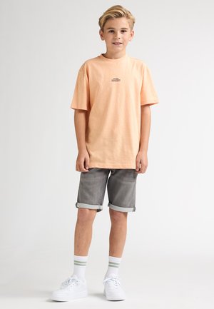 Camiseta de manga corta color durazno con logotipo negro, combinada con pantalones cortos grises remangados. El look se completa con zapatillas blancas y calcetas blancas con rayas verdes.