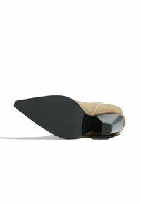 Zapato de tacón alto beige con diseño de punta afilada. La suela es negra y el tacón tiene un acabado plateado elegante con una superficie texturizada.