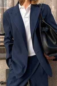 Blazer oversize bleu marine, chemise polo blanche, pantalon assorti et sac fourre-tout en cuir noir. Le tissu est lisse avec des épaules structurées.