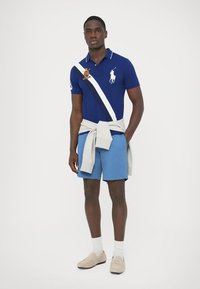 Tumši zila polo krekla ar baltām un tumši zila diagonālām svītrām, uzdrukāts ar zirga logotipu. Kombinēts ar gaiši zilām šortām un bēšām slip-on kurpēm.