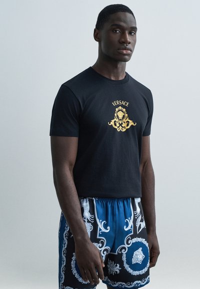 Schwarzes Baumwoll-T-Shirt mit einem goldfarbenen "Versace"-Emblem, kombiniert mit blau-schwarzen Badehosen mit weißen Akzenten und Details.
