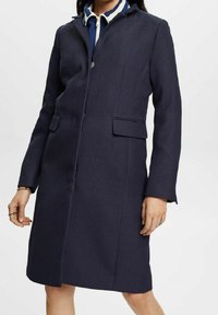 Cappotto lungo blu navy in tessuto testurizzato con vestibilità sartoriale, chiusura a bottoni nascosta e due tasche frontali. Presenta un colletto con incisione.