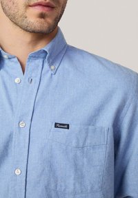Camisa de botones azul claro con cuello, botones blancos y un bolsillo en el pecho izquierdo que presenta un parche de logo negro. Tejido suave y texturizado.