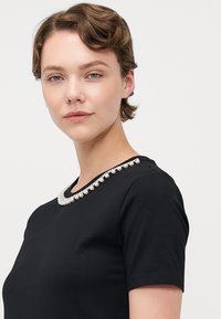 Svart t-skjorte med rund hals, pyntet med en rekke falske perler og rhinestones, korte ermer og en myk tekstur.