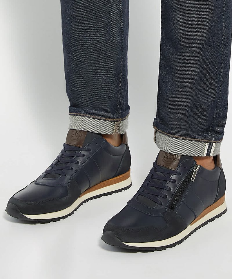 Marineblauwe sneakers met bruine accenten en ritsen aan de zijkant, gedragen met donkere opgerolde spijkerbroeken op een egale lichte achtergrond.