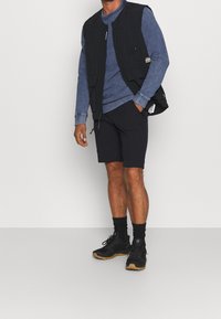 Musta ilmanhiha vest kumppanina siniselle pitkähihhaiselle paidalle, yhdistettynä tummiin shortseihin ja mustiin kenkiin, joissa on ruskeat pohjat. Rentoa, kerroksellista pukeutumista.