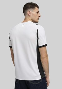 Camiseta deportiva blanca con acentos negros en los laterales y las mangas, con cuello redondo y un logotipo con "1901" en la parte posterior del cuello.