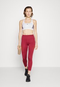 Biały stanik sportowy z czarnymi akcentami, wysokie bordowe legginsy i czarne buty sportowe. Trzyma beżową butelkę na wodę.