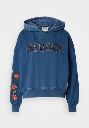 Zila kokvilnas džemperis ar kapuci, ar vārdu "HEROES" lieliem burtiem uz priekšpuses un ziedu izšuvumu uz viena piedurknes.