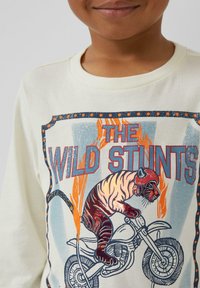 Kind trägt ein cremefarbenes Hemd mit einem grafischen Motiv eines Tigers in einem Helm, der auf einem Motorrad durch Flammen fährt, betitelt mit "DIE WILDEN STUNTS."