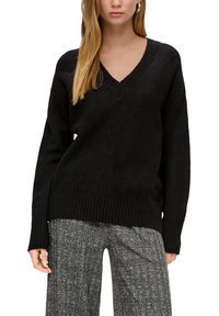 Schwarzer V-Ausschnitt-Pullover mit gerippter Textur und langen Ärmeln, mit einem lockeren Schnitt und geradem Saum. Kombiniert mit grauen gemusterten Hosen.