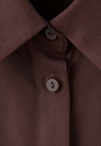 Gros plan d'un col de chemise marron foncé avec deux boutons assortis attachés sur un tissu texturé.